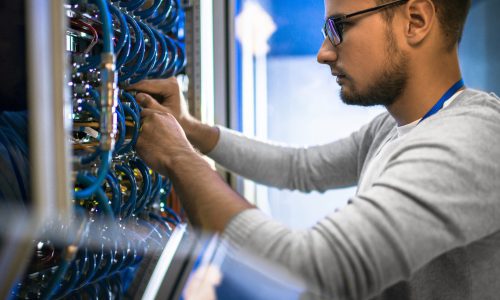 Side view portrait of young man wearing glasses connecting cables in server cabinet while working with supercomputer in data center